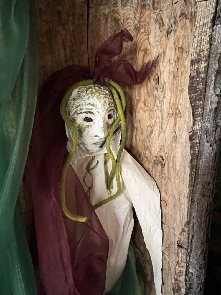 Maskenfigur mit grünem Haar und rotem Tuch vor altem Holz
